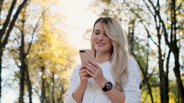 Портрет дівчини в білому светрі, що пише повідомлення на своєму смартфоні на відкритому повітрі. Жінка використовує цифровий гаджет, прокручує через соціальні мережі на пристрої, читає новини на додатку, на заході сонця . — стокове відео