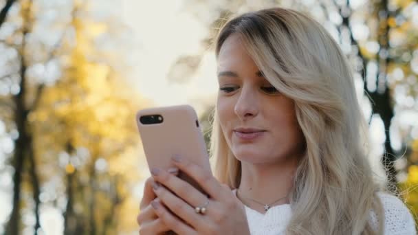 Close up portrait of cute girl wearing white sweater using smart phone and browsing on mobile phone in city park. Portrait of woman writing message on her smartphone at sunset. — Stock Video