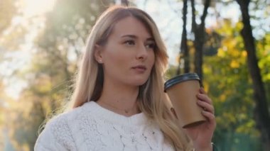 Kamera gitmek istimal telefon ve kahve içme genç çekici hipster sarışın kız izler ve park, yumuşak güneş ışığında etrafta dolaşıyor. Kadın kamera ve gülümseme bakarak. Portre kadar kapatın