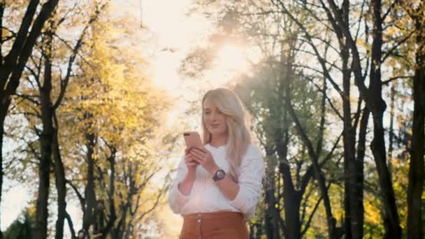 Menina sorridente bonito jovem elegante usando telefone inteligente moderno enquanto caminha no parque da cidade. Lady digitando mensagem de texto em rolos de celular através das mídias sociais, lendo notícias no aplicativo, ao pôr do sol . — Vídeo de Stock