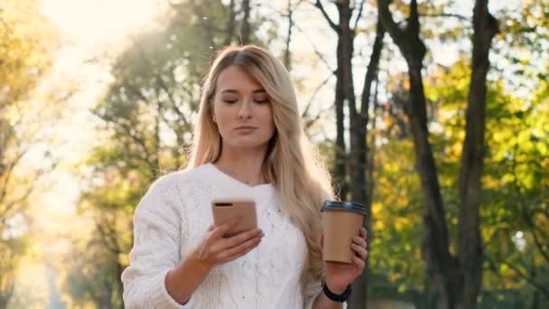 Menina sorridente bonito jovem elegante usando telefone inteligente moderno enquanto caminha no parque da cidade. Lady digitar mensagem de texto no celular e beber café para ir, ao ar livre, ao pôr do sol . — Vídeo de Stock