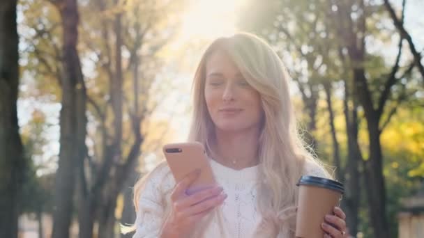 美丽的年轻嬉皮士女孩穿着白色毛衣使用现代智能手机 而在城市公园休息散步 女雇主在手机上打字短信外 慢动作 — 图库视频影像