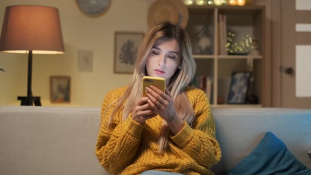 Mujer joven rubia bonita sentada en el sofá mirando sus aplicaciones en el teléfono inteligente en una habitación acogedora. Mirada relajante y reflexiva. Encantadora atmósfera casera . — Vídeo de stock