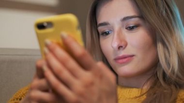 Güzel rahat ev odasında otururken sarı Smartphone'da taping güzel kız portresi kapatın. Kaydırma, dokunmatik ekran yazarak, app kullanarak ve browsing belgili tanımlık enterne kadın