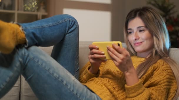 Ελκυστική ξανθιά φοιτητής κορίτσι γραπτά μηνύματα στο τηλέφωνό της. Γυναίκα που χρησιμοποιούν smartphone για να επικοινωνούν με τους φίλους στο κοινωνικό δίκτυο πληκτρολόγηση μηνυμάτων και να ψάχνετε μέσω του internet που κάθεται στον καναπέ άνεση στο σπίτι — Αρχείο Βίντεο