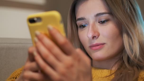 Primer plano retrato de la hermosa chica grabando en el teléfono inteligente amarillo mientras está sentado en la acogedora sala de estar. Mujer desplazándose, escribiendo en la pantalla táctil, utilizando la aplicación y la navegación en Internet — Vídeos de Stock