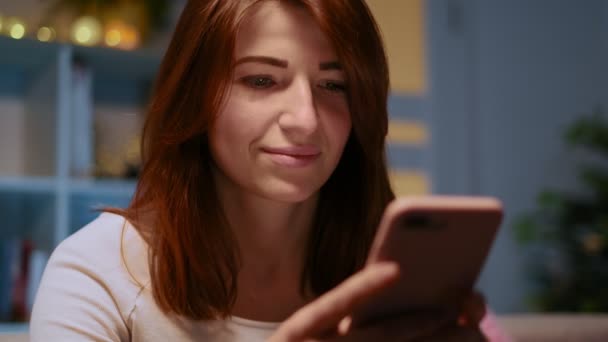 Mensajería de mujer joven en el teléfono inteligente en casa, sentado cómodamente en el sofá beige por la noche. Chica atractiva mirando y sonriendo mientras envía mensajes de texto en el teléfono móvil, utilizando la aplicación. Retrato de primer plano . — Vídeos de Stock