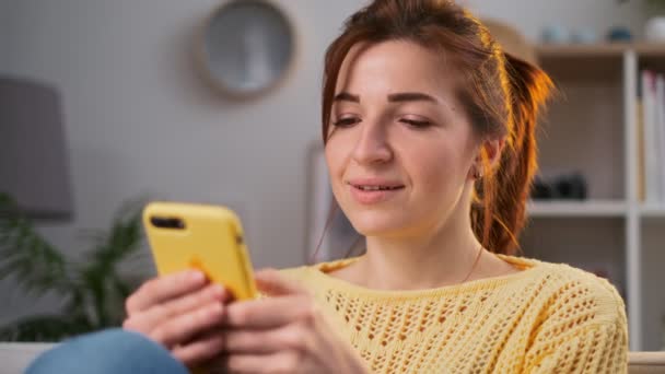 Hermosa mujer joven en el suéter amarillo sosteniendo un teléfono inteligente en las manos y escribiendo en él utilizando la aplicación, charlando en la sala de estar en casa. Encantadora atmósfera casera — Vídeos de Stock