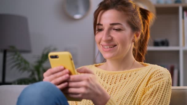 Portret van jonge alleenstaande vrouw met behulp van dating app op haar smartphone. Meisje in de gele trui een smartphone in handen te houden en te typen op het babbelend in de woonkamer. Mooie huis sfeer. — Stockvideo