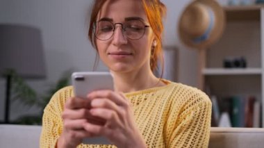 Kız bardaklarda evde oturma odasında kanepede oturan smartphone cep telefonu ile mesaj yazmaya konsantre. Kapalı. Portre. Çekici bir kadın onun beyaz Smartphone'da kaydırma. Yakın çekim. Kapalı.