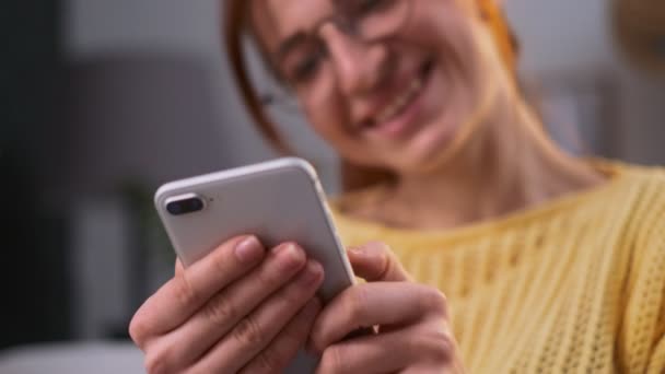 Primer plano retrato de mujer joven caucásica con gafas sentarse en el sofá, utilizando el teléfono inteligente en casa. Chica concentrada en gafas mensaje de escritura, en la sala de estar, desplazándose en su teléfono inteligente blanco . — Vídeos de Stock