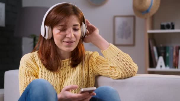 Girl singing song, young caucasian woman relaxing, sitting on sofa and listening to music. Leisure and relaxation at home. Beautiful woman listening to music at trendy apartment with headphones. — Stock Video