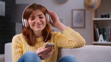 Kız şarkı şarkı, rahatlatıcı, kanepede oturan ve evde müzik genç Kafkas kadın portresi. Güzel kadın trendy daire kulaklık ile müzik dinlemek.