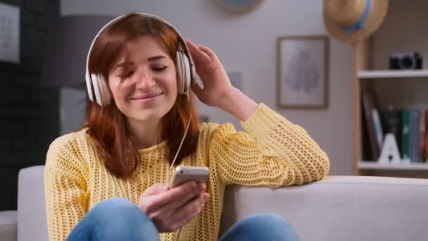 Portrait of girl singing song, young caucasian woman relaxing, sitting on sofa and listening to music at home. Beautiful woman listening to music at trendy apartment with headphones. — Stock Video