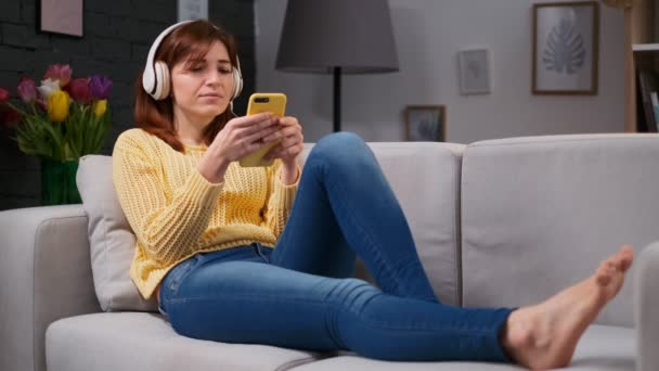 Retrato de una linda chica en auriculares tumbados en un sofá en la sala de estar escuchando música y navegando en el teléfono móvil en casa. Mirada relajante y reflexiva. Encantadora atmósfera casera . — Vídeos de Stock