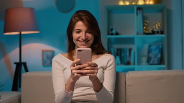 Portret van de jonge vrouw zittend op de Bank chatten op de mobiele telefoon en lachen, in een gezellige kamer. Binnen. Meisje met app. mooi huis sfeer. — Stockvideo