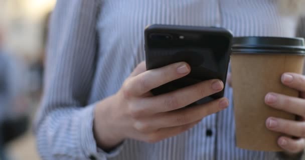 Wanita bisnis muda dalam kemeja memegang ponsel dan kopi di tangannya dan SMS, pandangan close-up . — Stok Video