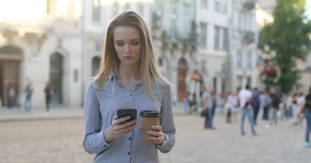 Mujer de negocios bastante joven mensajes de texto en su teléfono en la calle con café en la mano . — Vídeo de stock