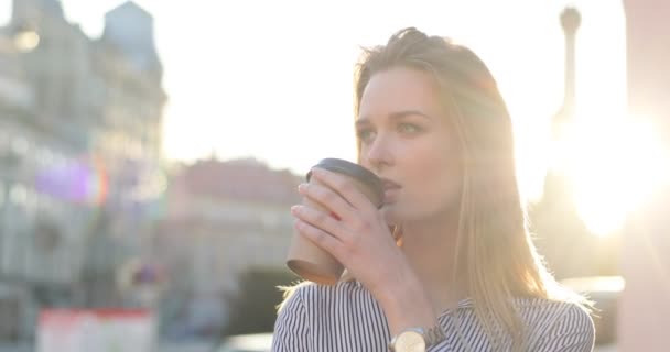 Una bella donna bionda di successo in camicia sta bevendo caffè da asporto all'alba in città, ritratto da vicino, vista laterale. Il sole sta lampeggiando la telecamera — Video Stock