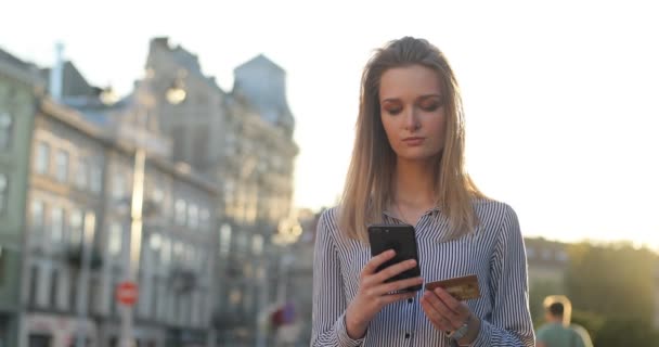 Una mujer rubia con estilo está ingresando un número de tarjeta de crédito en su teléfono en la calle y sonriendo a la cámara. Banca móvil . — Vídeos de Stock