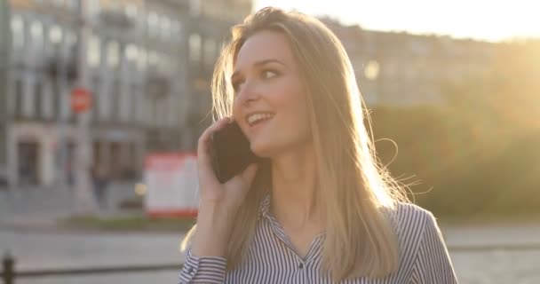 Close-up portret van een vrolijke dame praten aan de telefoon en lopend onderaan de straat met een glimlach op haar gezicht, zonlicht in de camera. — Stockvideo