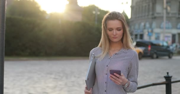 En attraktiv blond klädd i en tröja håller en anteckningsbok och textning på sin telefon. Sedan berör hon sitt hår och leenden till kameran på gatan. Framifrån. — Stockvideo