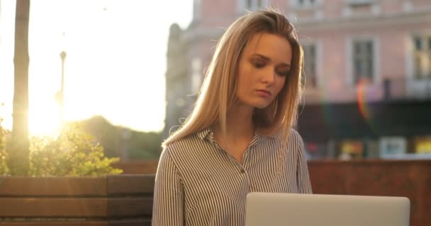 Uma jovem senhora de negócios loira está sentada em um banco em um dia ensolarado e trabalhando em seu laptop, close-up . — Vídeo de Stock