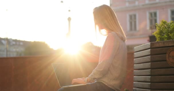 Vista lateral de uma linda garota loira sentada em um banco e trabalhando em seu laptop ao pôr do sol . — Vídeo de Stock