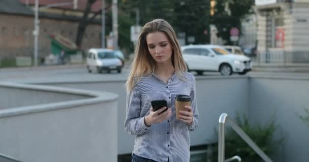 魅力的な若い女性がタイプしている間に彼女の手でコーヒーと電話通りを歩いてください。. — ストック動画