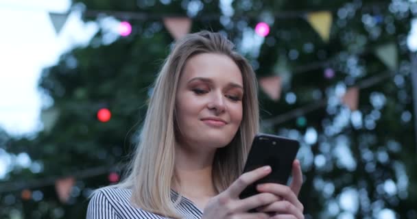 Een close-up portret van een mooie Kaukasische meisje met heldere ogen typen op haar mobiele telefoon en glimlachend buitenshuis. Vooraanzicht. — Stockvideo