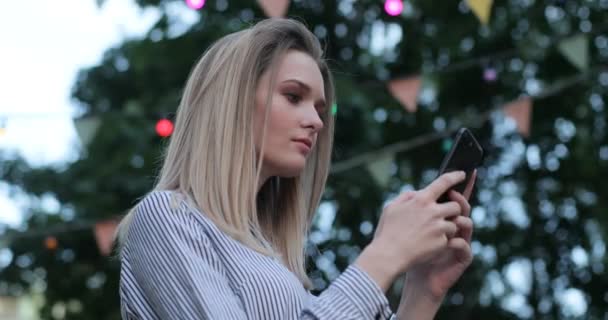 Una hermosa chica está escribiendo en su teléfono y sonriendo alegremente al aire libre. Vista lateral, retrato de primer plano . — Vídeos de Stock