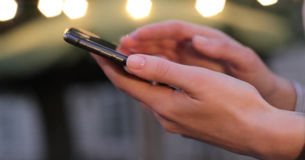 Delicate vrouwelijke handen scrollen iets op mobiele telefoon. 'S avonds lichten op de achtergrond. Close-up. — Stockvideo
