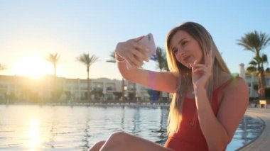 Sarışın kadın kırmızı mayo portre çekim selfie akıllı telefon ile lüks bir Villa Yüzme Havuz kenarında rahatlarken yapıyor. O poz, hareketli ve dikkatsizce gülümseyen.