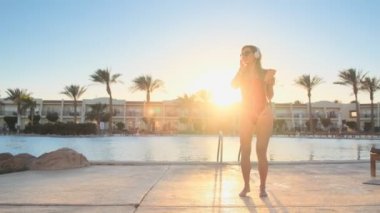Çekici neşeli kız kırmızı mayo ve kulaklık, eğlenceli dans ve Yüzme Havuz Partisi yakınındaki smartphone cep telefonu ile müzik dinlemek. Bikini kadın güneş gözlüğü yaz tatilde iyi vakit geçiriyor.