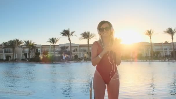 Mooie vrouw in rode zwembroek en koptelefoon, dansen en muziek op smartphone luisteren. Portret van bikini girl in zonnebrillen hebben goede tijd, ontspannen in de buurt van het zwembad in luxevilla. — Stockvideo