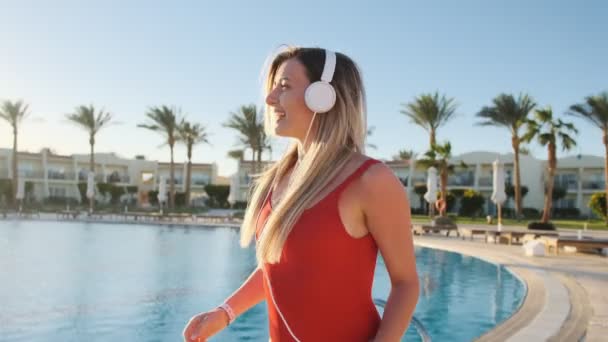 Gyönyörű nő piros fürdőruha és fejhallgató tánc közelében világos háttér kék medence, szórakoztató és hallgat zenét a smartphone segítségével app. Bikini lány a nyári jó idő. — Stock videók