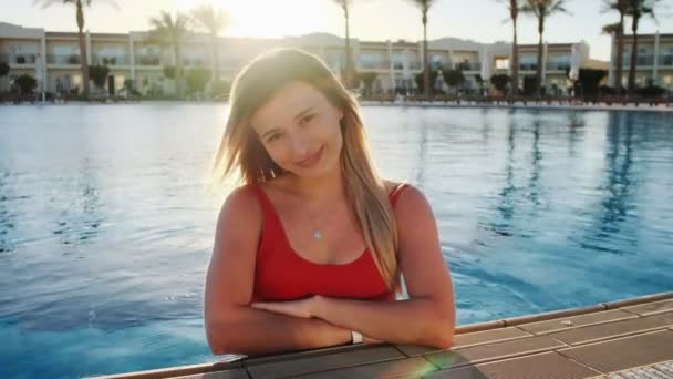 微笑的年轻女孩的肖像, 看着相机。美丽的女人穿着红色的泳衣坐在蓝色的游泳池在炎热的阳光明媚的日子。比基尼女孩玩得开心, 暑假在豪华酒店 — 图库视频影像
