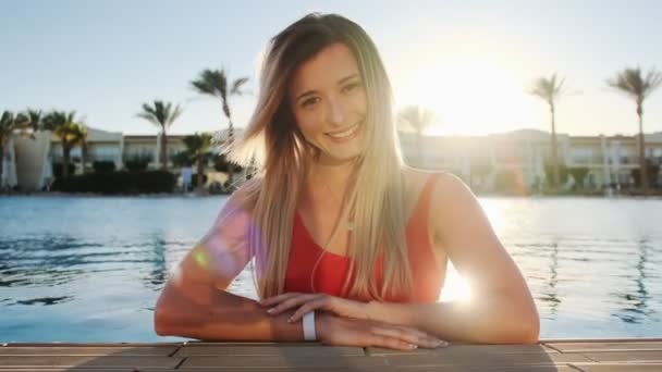 Portrét s úsměvem dívka, při pohledu na fotoaparát. Krásná žena v červených plavkách sedí v modré bazénu na horký slunečný den. Bikini holka dobře bavíte, letní dovolenou v luxusní vile — Stock video