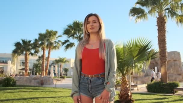 Retrato de uma jovem hipster sorridente, a olhar para a câmara. Mulher bonita em maiô corpo vermelho no dia ensolarado quente. Férias de verão no hotel de luxo. Piscina e palmas de fundo . — Vídeo de Stock