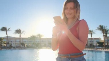 Havuz tarafı mavi su arka plan ile yakın kız gülümseyerek, o bir akıllı telefon app, mesaj kullanarak. Seksi kadın kırmızı moda vücut mayo lüks otelde cep telefonuna ileti.