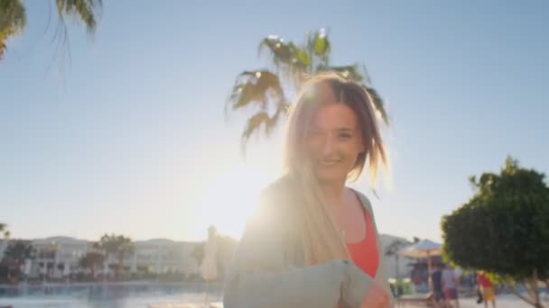 Sígueme, joven chica corriendo riéndose pidiéndote que la sigas en vacaciones. Mujer en traje de baño rojo saltando, en un día caluroso y soleado. Vacaciones de verano en hotel de lujo, piscina de fondo y palmas — Vídeo de stock