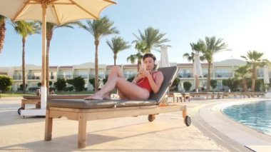 Kırmızı bikini beyaz kulaklık otel havuz yan sandalyede güverte üzerinde yatarken, onun smartphone üzerinden müzik dinleme çekici genç kadın. Güneşlenme ve rahatlatıcı resort adlı bir kız.