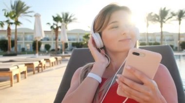 Kırmızı bikini beyaz kulaklık otel havuz yan sandalyede güverte üzerinde yatarken, onun smartphone üzerinden müzik dinleme gülümseyen genç çekici genç kadın portresi. Kız rahatlatıcı Resort.