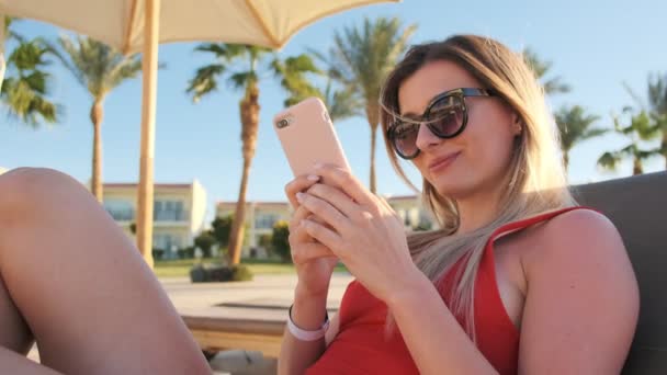 Portret van lachende aantrekkelijk meisje in een rode bikini met haar smartphone in hotel zwembad zijde. Vakantie reizen vrouw ontspannen bij spa resort, Oceaan, zee en strand. — Stockvideo