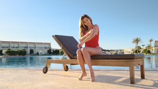Привлекательная девушка в красных купальниках наносит лосьон для тела на чувствительную кожу, чтобы защитить кожу и предотвратить старение от солнца. Загорание на экзотическом курорте рядом с бассейном . — стоковое видео