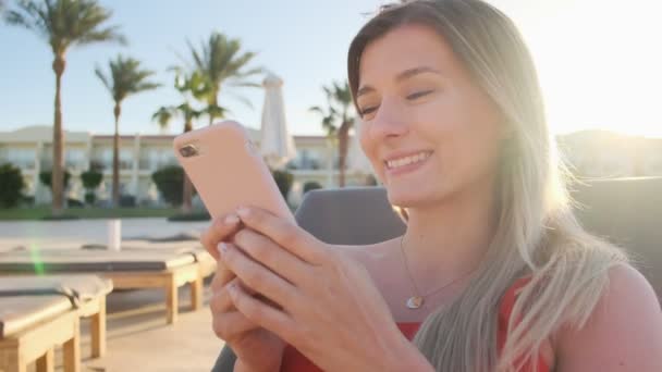 Retrato de una mujer descansando en una tumbona usando un teléfono inteligente, navegando por Internet, se desplaza a través de las redes sociales. Joven chica feliz con bikini rojo usando el teléfono móvil mientras toma el sol en la piscina . — Vídeos de Stock