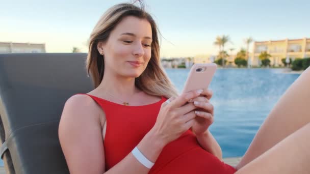 Γυναίκα που χρησιμοποιεί κινητό στο ανάκλιντρο στην παραλία. Καλοκαίρι κορίτσι με κόκκινο μπικίνι περιήγηση στο διαδίκτυο, μηνύματα και κύλιση στα νέα της στο smartphone, ηλιοθεραπεία στην πισίνα. — Αρχείο Βίντεο