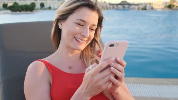 Mujer bikini sonriente retrato usando teléfono móvil en chaise long. Chica de verano en bikini rojo navegar por Internet, mensajería y se desplaza a través de sus noticias en el teléfono inteligente, tomar el sol en la piscina . — Vídeo de stock