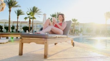 Havuz tarafı Plaj yakın güverte sandalyede yatarken güzel genç kadın akıllı telefonda internet tarama yan görünümü. Rahat kız manifatura mesaj ve tatil cep telefonu ile bloglama.