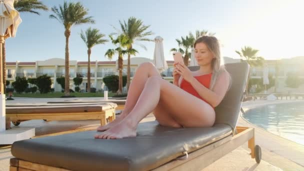Girl resting on a deck chair using smartphone, browsing the internet, scrolls through social media. Young woman wearing red bikini using mobile phone while sunbathing by the pool. — Stock Video